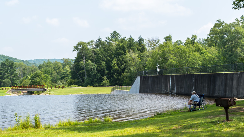 Man in a white shirt fishing by a lake, with a grill to his right and a cement dam behind him, surrounded by trees with mountains in the background, under a blue sky