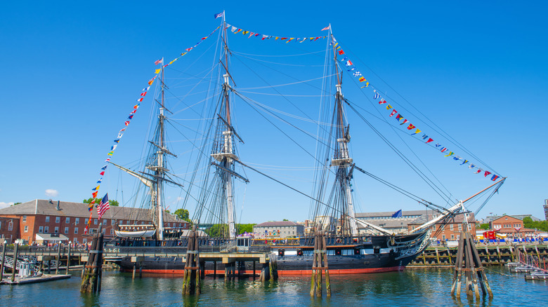 The USS Constitution floats in the harbor in Boston, MA, in front of a blue sky and red brick buildings