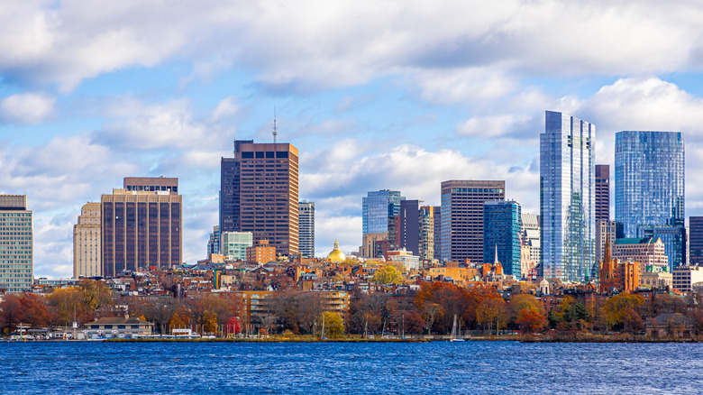 Boston's skyline in front of a cloudy blue sky and a dark blue Atlantic with fall colors adorning the trees