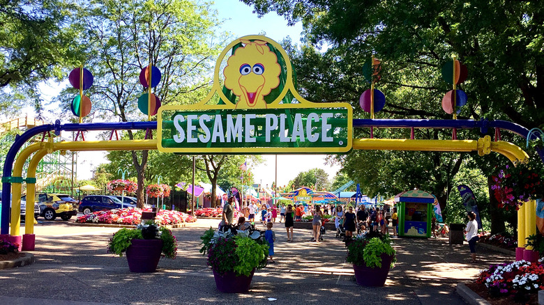 Sesame Place sign with a roller coast, rides, and crowds in the background