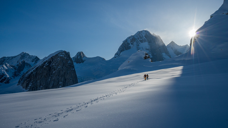 Two people backcountry skiing outside of Sheldon Chalet, a five-star luxury lodge located on a nunatak in Denali National Park, Alaska, with the sun peaking out from behind a mountain.