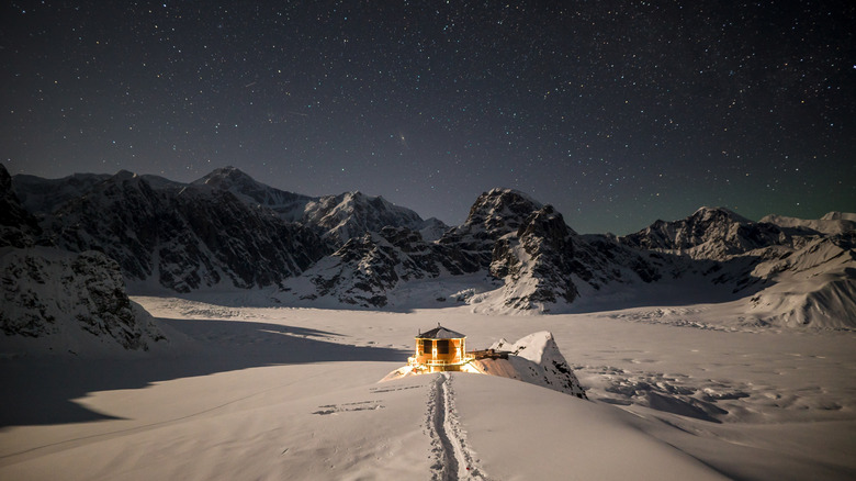 Aerial view of the exterior of Sheldon Chalet, a five-star luxury lodge located on a nunatak in Denali National Park, Alaska, surrounded by snow-covered peaks with stars twinkling in the night sky in the background.