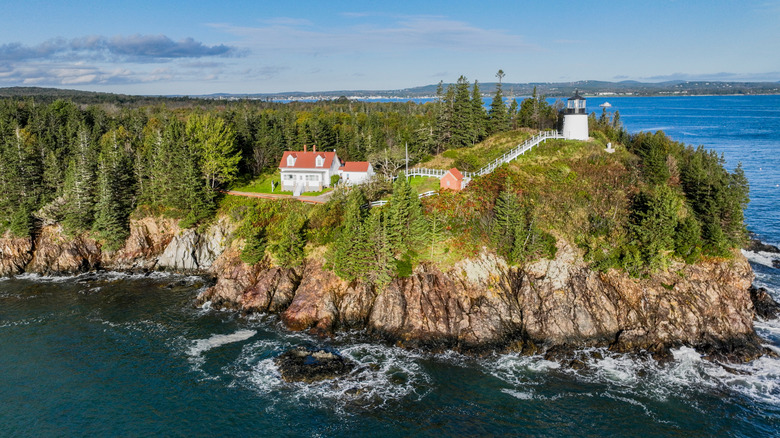 Owl's Head Lighthouse near Rockland, Maine, with green trees, a rocky shore lapped with white water waves, a dark blue sea, and a sky with distant stratus clouds and even more distant cirrostratus clouds