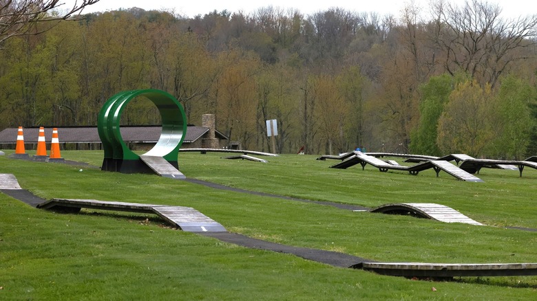 Children's Bike Park at Cross Creek Park, with wooden ramps, a green tube, and orange traffic cones surrounded by grass, with a pavilion and winter trees in the background