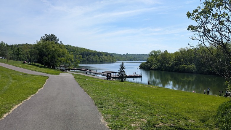 Cross Creek Lake, with a paved trail through grass leading to wooden docks along the water lined in trees, with people fishing from the shore on the right side of the frame, under a cloudy blue sky