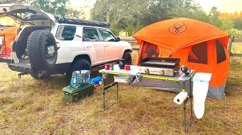 An outdoor camping setup features the Core FlexRail Table in front of an orange tent and white SUV