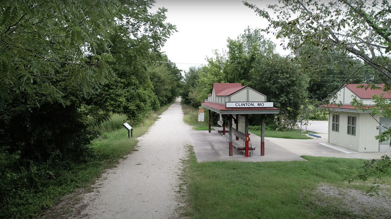 Green grass and green trees with a Clinton, MO sign above a bench on Katy Trail