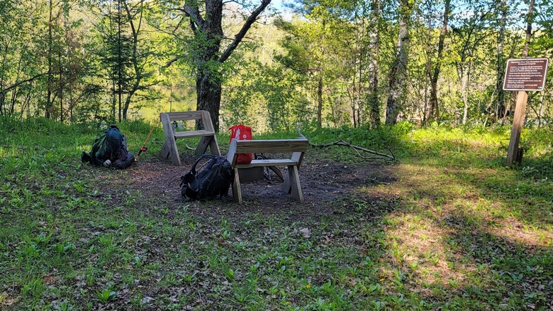 Primitive campsite at the Menominee River State Recreation Area, Wisconsin-Michigan border, with two benches, backpacks, a fishing rod, and a sign