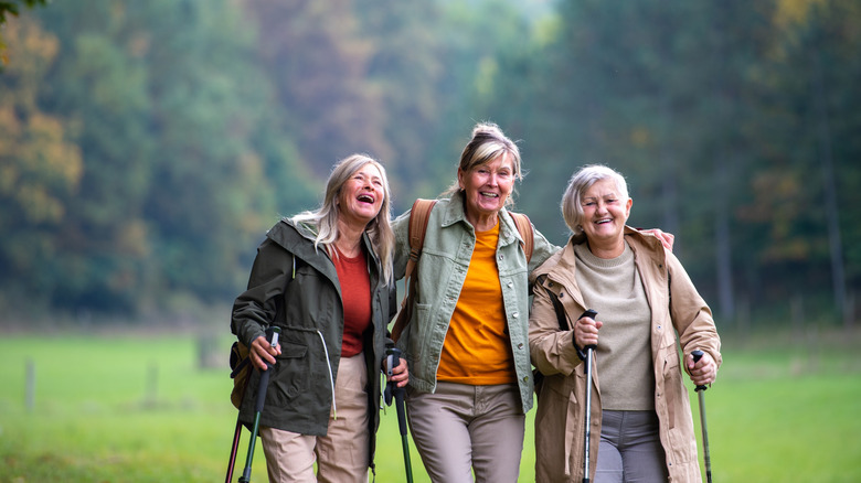 Three senior women smile while walking together through a field with trekking poles