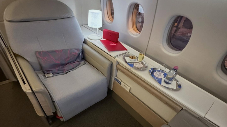View of the armchair, along with a lamp, menu, amenity kit, champagne, warm nuts, and water, in Air France's La Première Business Class
