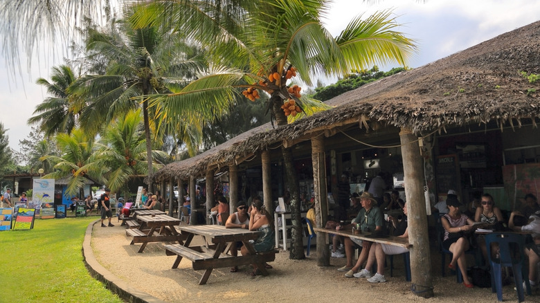 Tourists sitting on wooden benches under a thatched roof cafe under the shade of palm trees