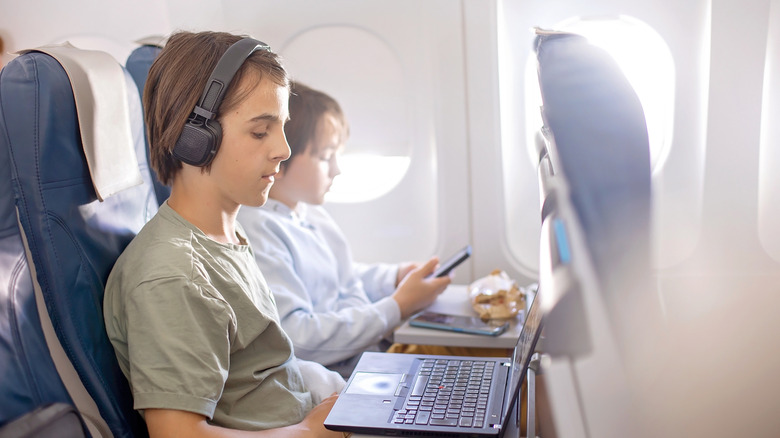 Two children, one watching a laptop and the other looking at a cell phone, on an airplane