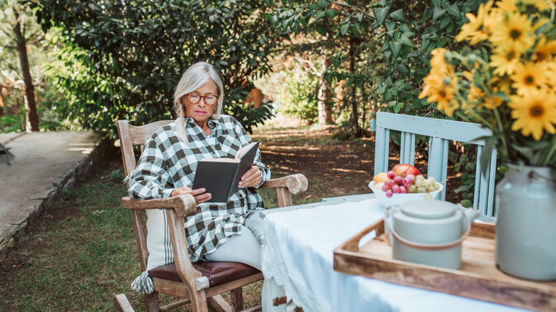 Woman reading a book in a rocking chair in a lush yard with trees and flowers