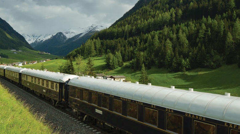 Exterior view of the Venice Simplon-Orient-Express, a luxury restored train from the 1920s, passing through a lush green valley with snowcapped mountains in the distance, somewhere in Europe