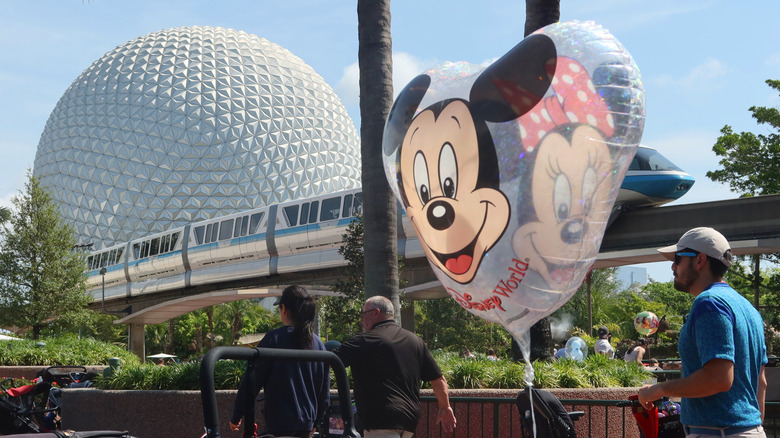 A monorail train passes behind a Mickey Mouse balloon on the grounds of Epcot theme park at Walt Disney World Resort in Orlando, Florida, on a bright day, as three visitors walk by
