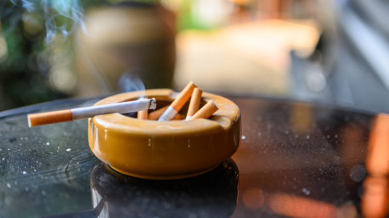A still-burning cigarette sits on the edge of a ceramic ashtray outside on a table, on a bright day.