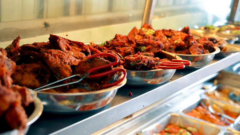 Steel dishes of spiced chicken with serving tongs behind a glass case