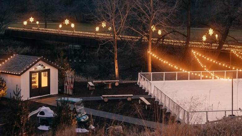 Small cabin with benches and an ice skating rink the foreground, with a road under lighted snowflake arches in the background
