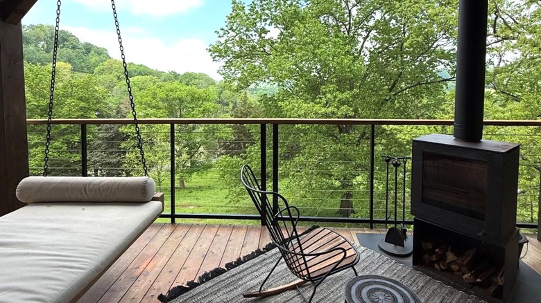 A timber deck with a swinging seat, chair, fireplace and view of green trees.