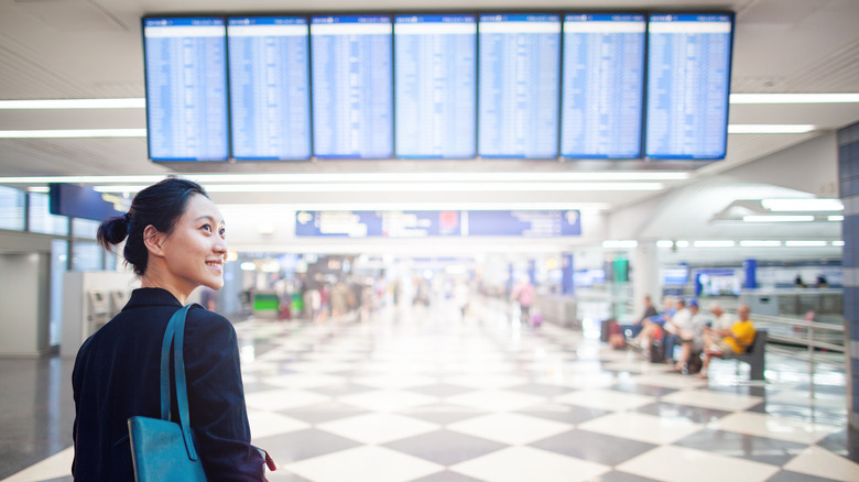 woman smiles while standing in front of flight departure screen at airport