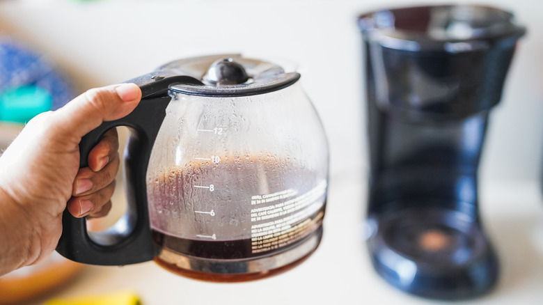 A hand holds a partially filled coffee carafe in front of a drip coffee machine in a hotel.