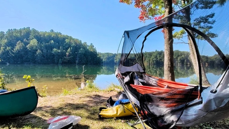 A green canoe and a camping hammock the shore of a calm lake with lush green trees in the background under a clear blue sky.
