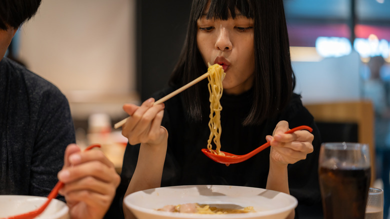 An Asian woman eating ramen with chopsticks and a red spoon