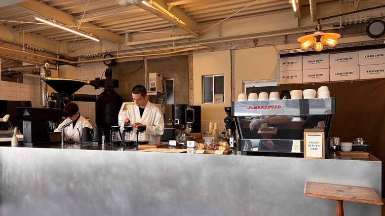 Two baristas preparing coffee in an aluminium-topped counter next to large coffee roasters and machines