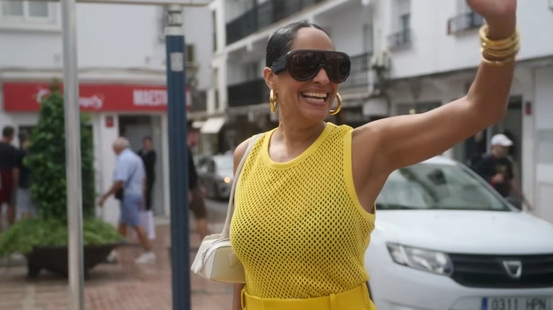 Tracee Ellis Ross waving on a street while wearing sunglasses and a yellow outfit