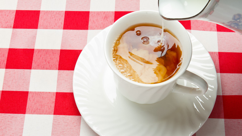 Milk mixing with hot tea in a white teacup on a saucer on a red checked gingham table cloth.