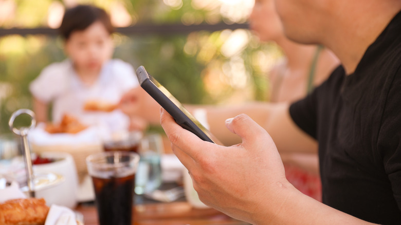 Man holding his phone at a dinner table. The other people in the background are blurred out.