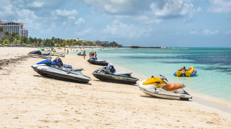 Jet skis sitting on a white-sand beach in Nassau with people, blue ocean, and hotels in the background.