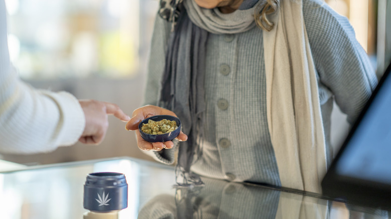 A person standing in a dispensary holds some marijuana buds in a measuring cup as another person's finger points at it
