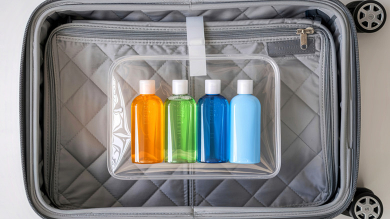 A clear zippered bag containing four colorful bottles of liquids is neatly placed inside an open suitcase, ready for travel.