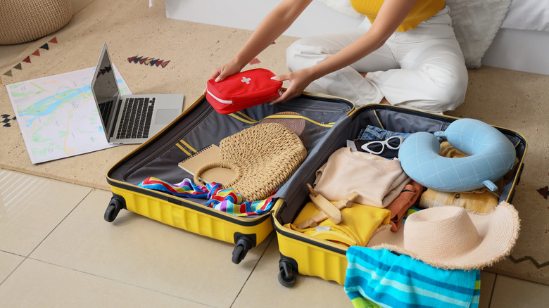 Young woman packing first aid kit into full suitcase already containing a neck pillow, sunhat, purse, and other items in bedroom