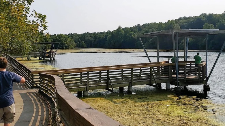 People fishing from a covered fishing pier at Valley Water Mill Park in Springfield, MO, featuring a calm lake, a paved pathway, and greenery.