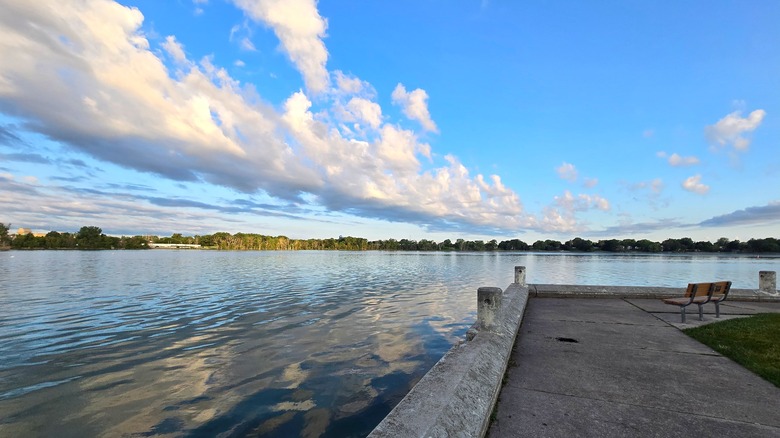 A bench on a path overlooks a calm river; trees on the opposite bank; and a blue, cloudy sky