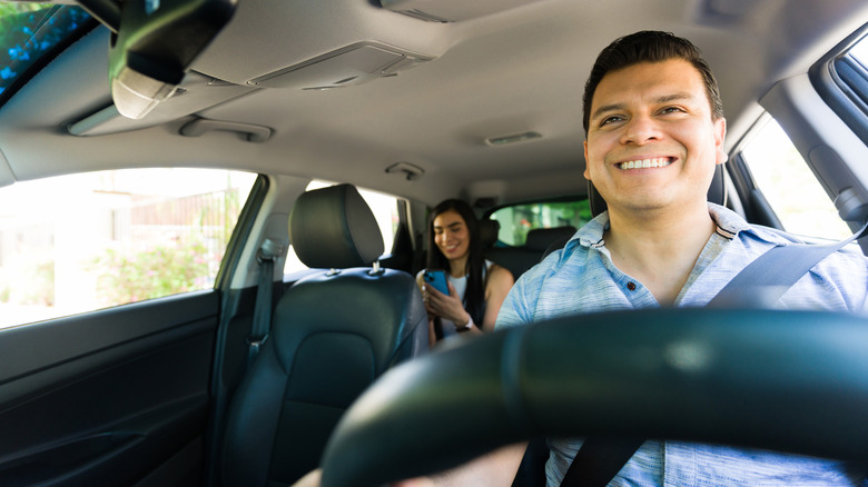 A man smiles from the driver's seat as his passenger in the back seat smiles at her phone