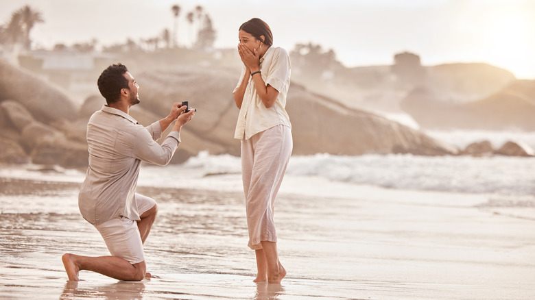 Man proposes to his girlfriend on beach at sunset, while she stares with emotion and surprise
