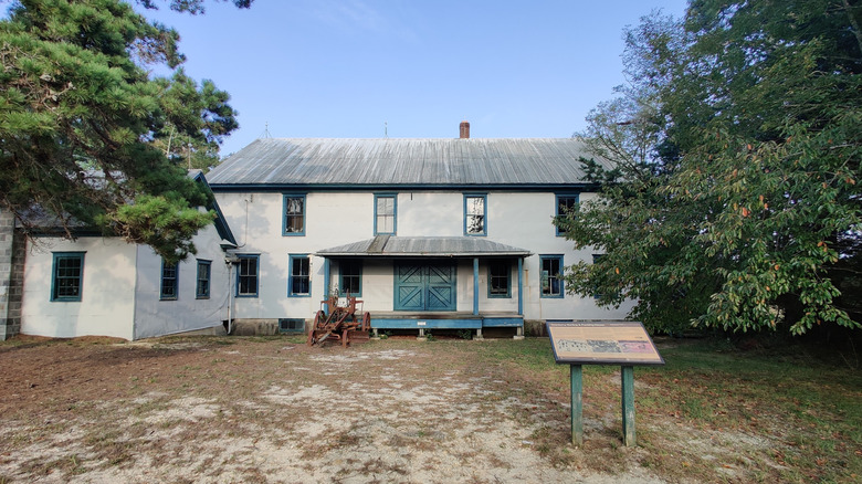 Double Tree Village Sorting and Packing House, a white two-story building with a blue door and gray roof, with red farm equipment and an interpretive sign out front and trees to either side, under a blue sky