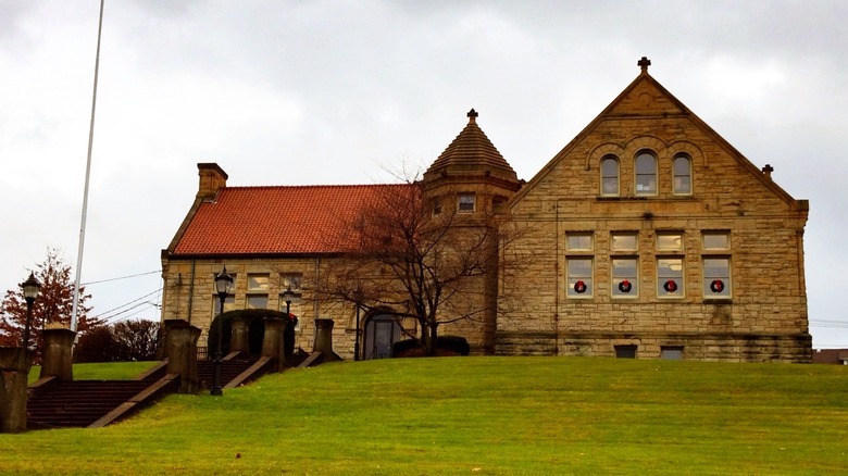 Carnegie Library of McKeesport, a brown stone building with a red roof and octagonal tower, with wreaths in four of the windows and a stone staircase leading to it through green grass, under a cloudy sky