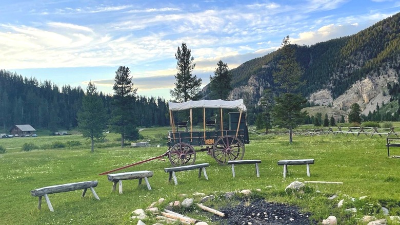 View of fire pit and wooden benches in foreground and historic carriage behind them, with green grass and hills with pines and a blue, cloud-dotted sky