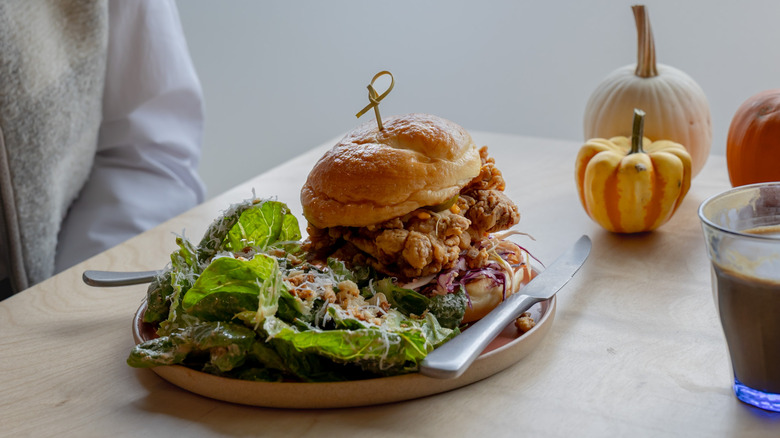 A plate with a fried chicken sandwich and salad sits on a table with miniature gourds in the backdrop and a drink in the foreground