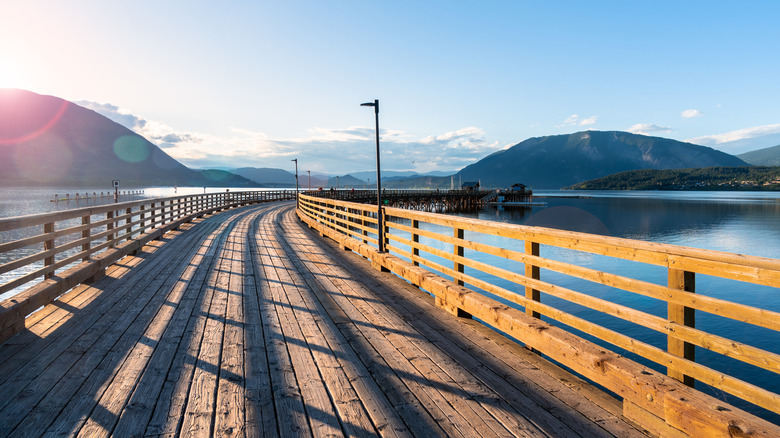 A wooden bridge arches out into a lake, with mountains in the backdrop