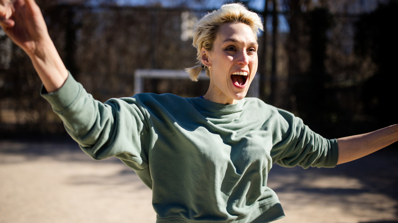 A woman with blonde hair exuberantly celebrating with outstretched arms outside