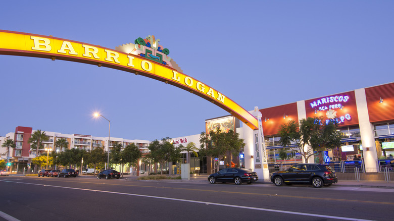 The illuminated Barrio Logan arch sign with trees and storefronts.
