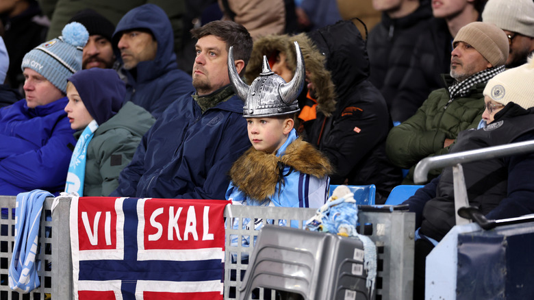 Football supporters in front of a Norway flag that reads "VISKAL," behind which a boy is wearning a horned helmet