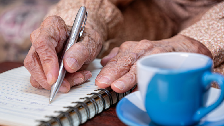 Closeup of wrinkled hands writing in a notebook with a coffee cup nearby