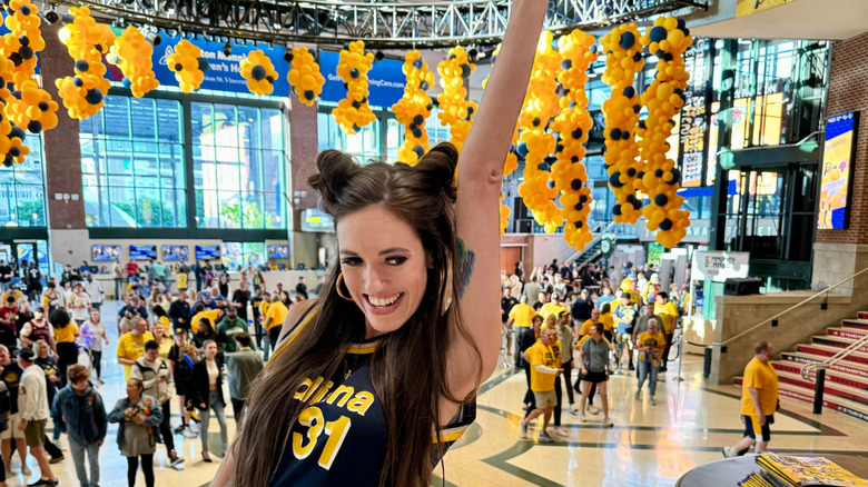 Brunette woman wearing a Reggie Miller 31 jersey posing in the lobby of Gainbridge Fieldhouse before a Pacers playoff game with a sea of other fans and balloons in the background