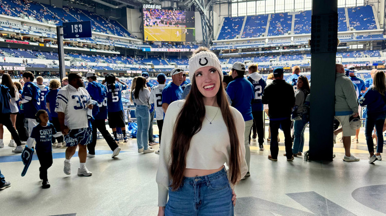Brunette woman wearing white sweater and Colts beanie with jeans stands inside of Lucas Oil Stadium on a Colts gameday with a crowd in the background